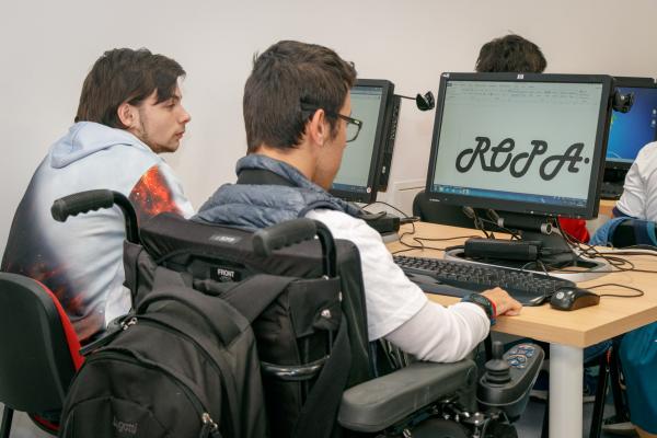 Dos alumnos jóvenes con discapacidad, uno en silla de ruedas, mirando las pantallas de sus ordenadores en un aula