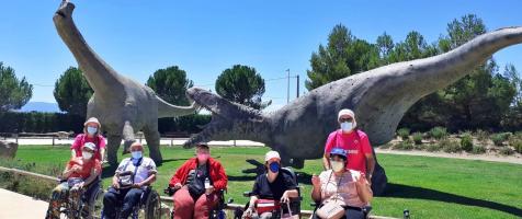 Cinco personas en silla de ruedas y dos voluntarios con camisetas rosas de la fundación posan ante las esculturas de dos dinosaurios gigantes