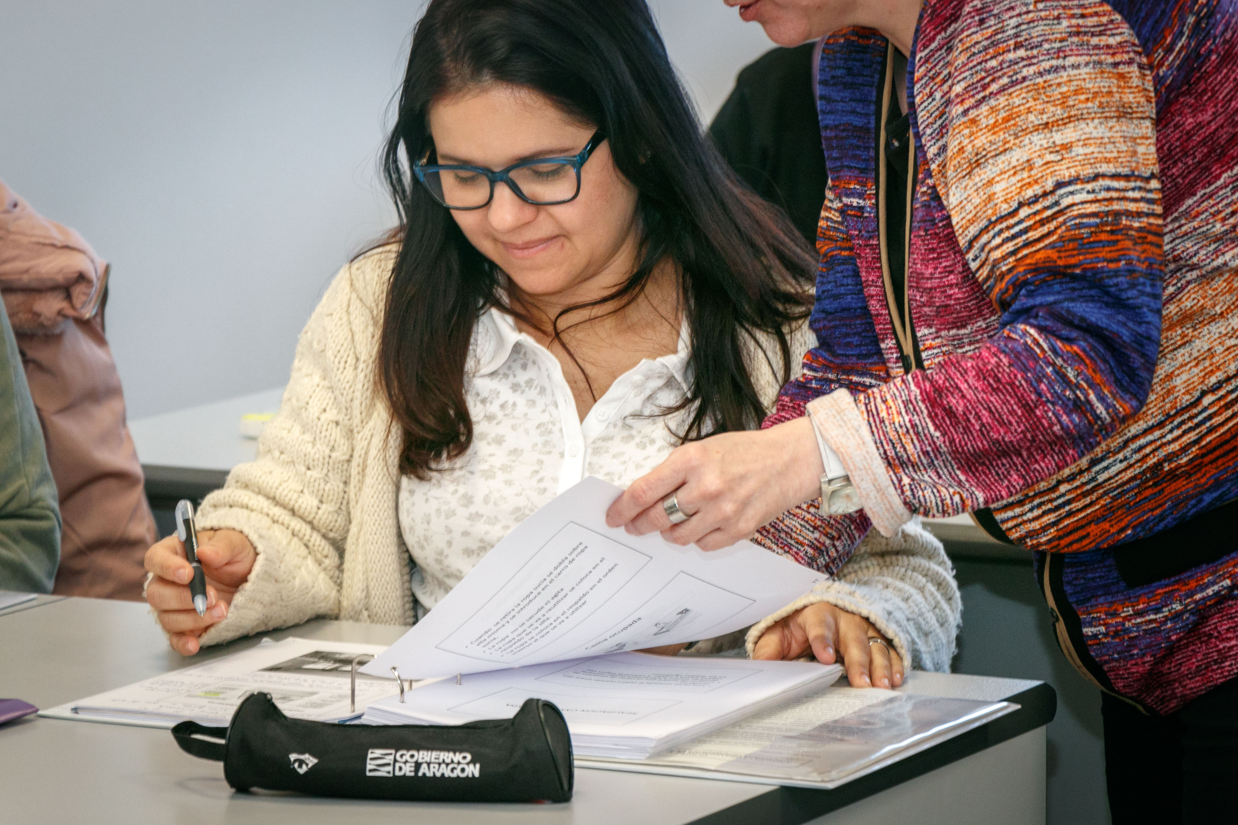 Mujer en un aula de formación sentada en una mesa mirando unos papeles que sujeta la profesora