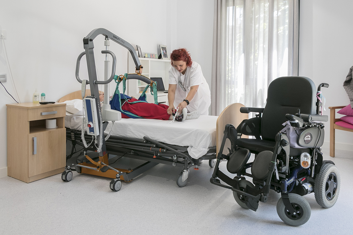 Habitación de residencia donde una auxiliar ayuda con una grúa a una mujer con discapacidad tumbada en una cama. A su lado hay una silla de ruedas automática 