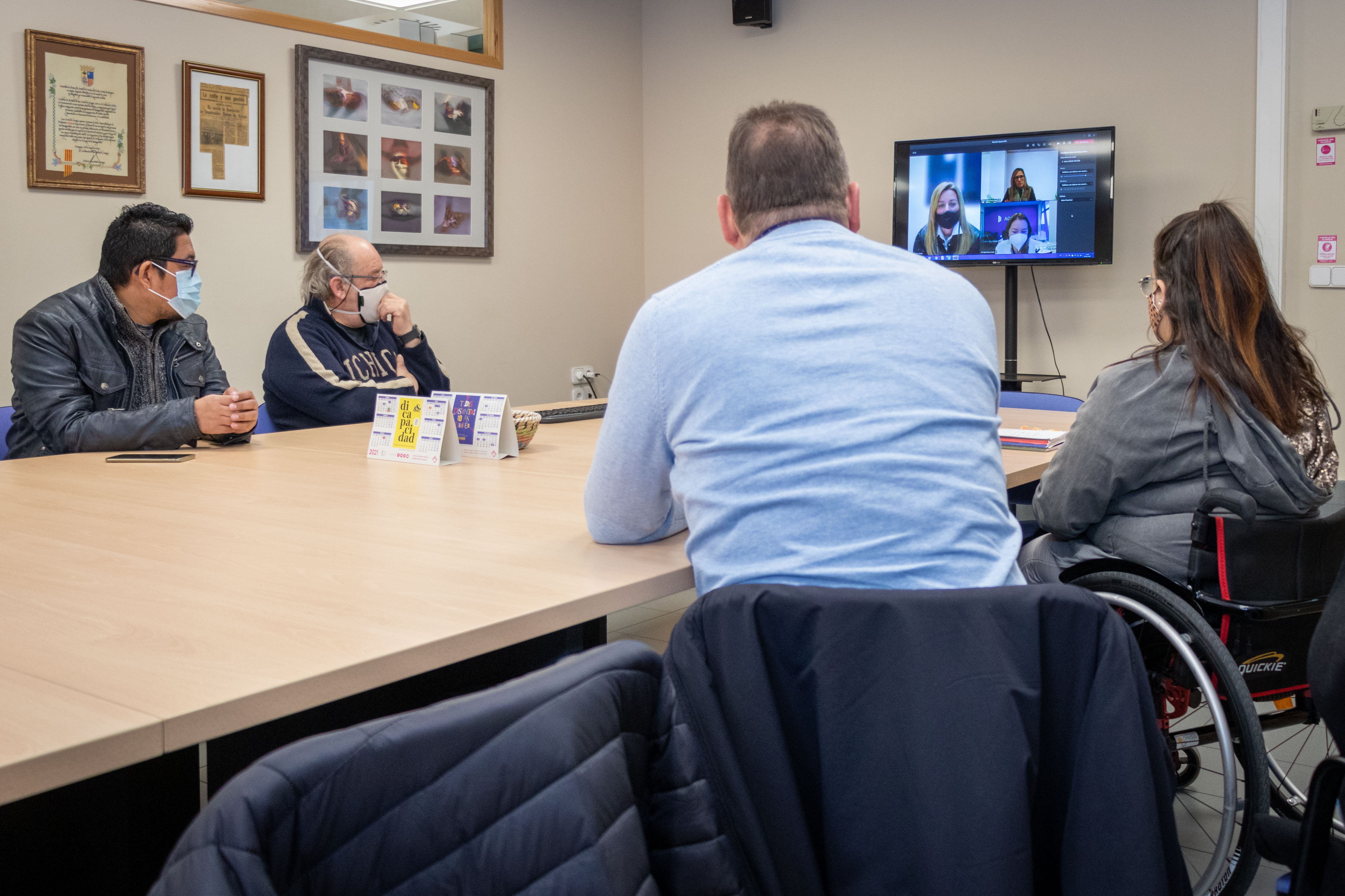Cuatro personas en una sala de reuniones sentadas en una mesa grande con una pantalla de televisión donde hay una videollamada