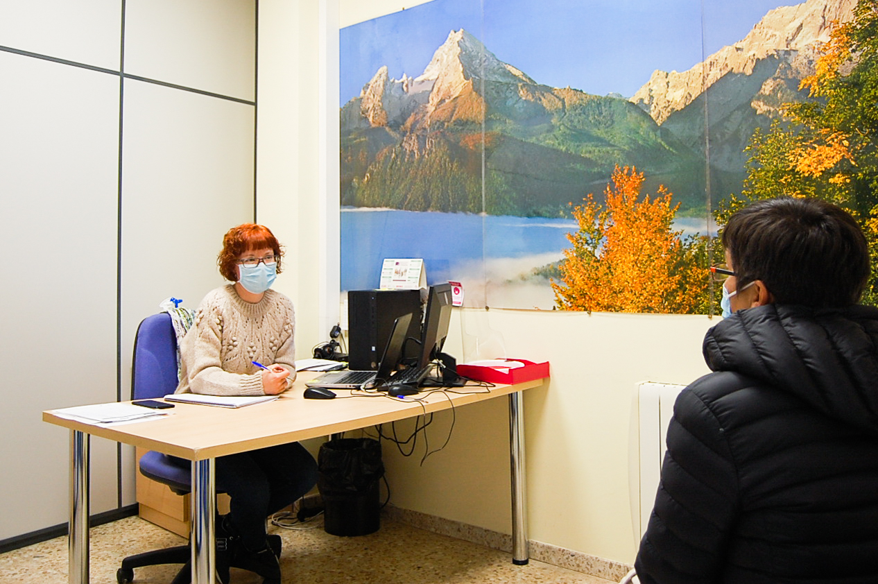 Una trabajadora social atiende a una usuaria en un despacho con una mesa y un cuadro de un paisaje