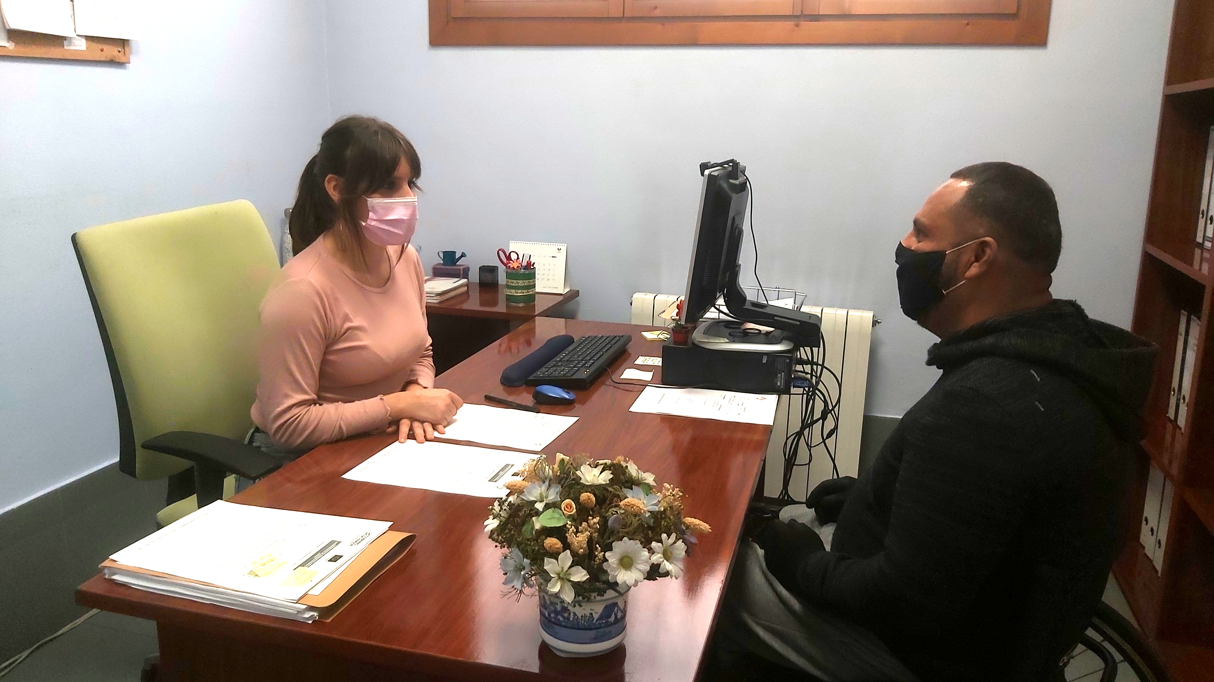 Una trabajadora social atiende a un usuario, ambos con mascarilla, en un despacho con una mesa y sillas