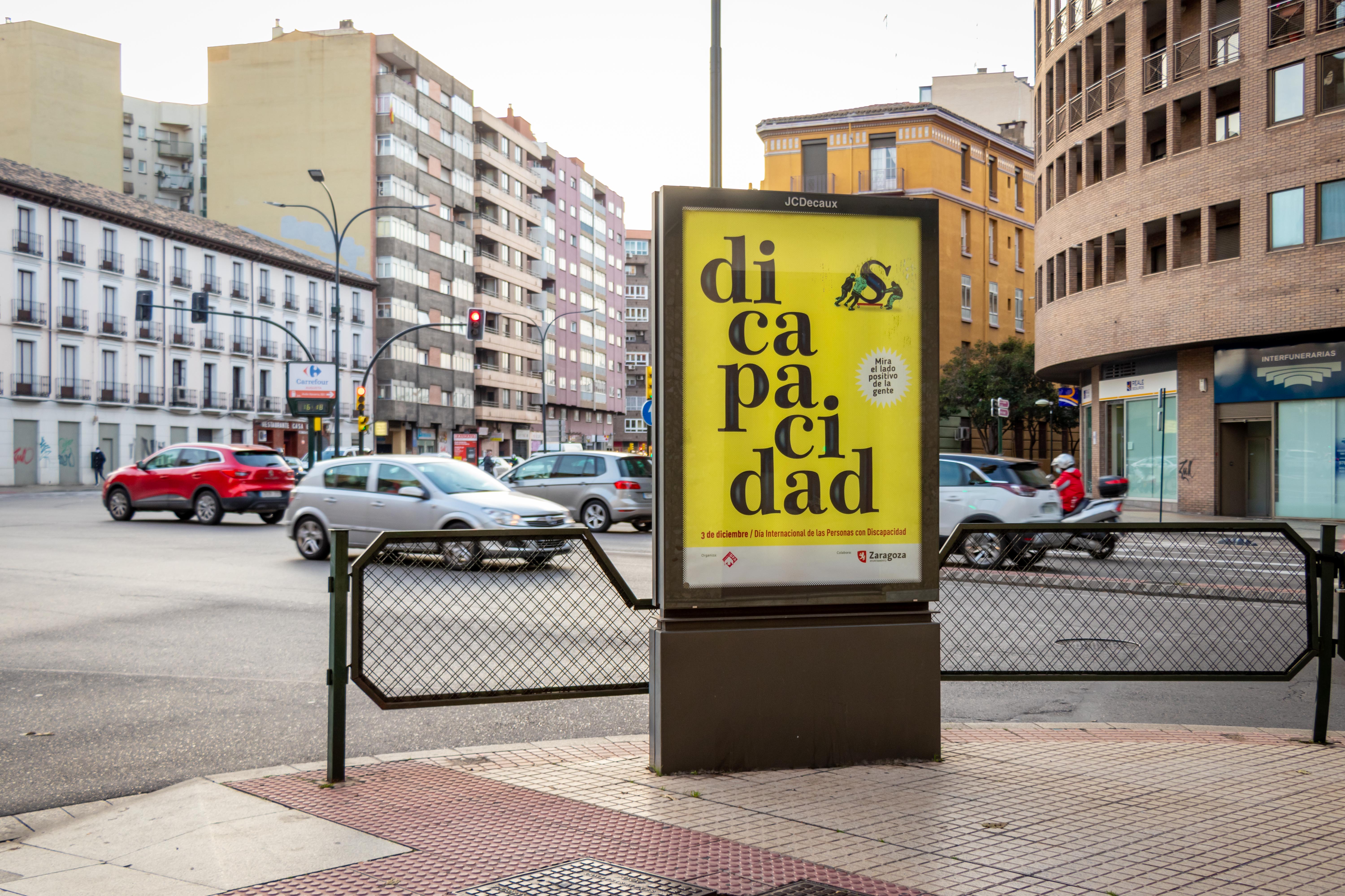 Calle de la ciudad con un anuncio publicitario con la campaña del 3 de diciembre de DFA: un cartel diseñado con fondo amarillo con un juego de palabras sobre discapacidad donde se lee Di capacidad mientras se aleja la S