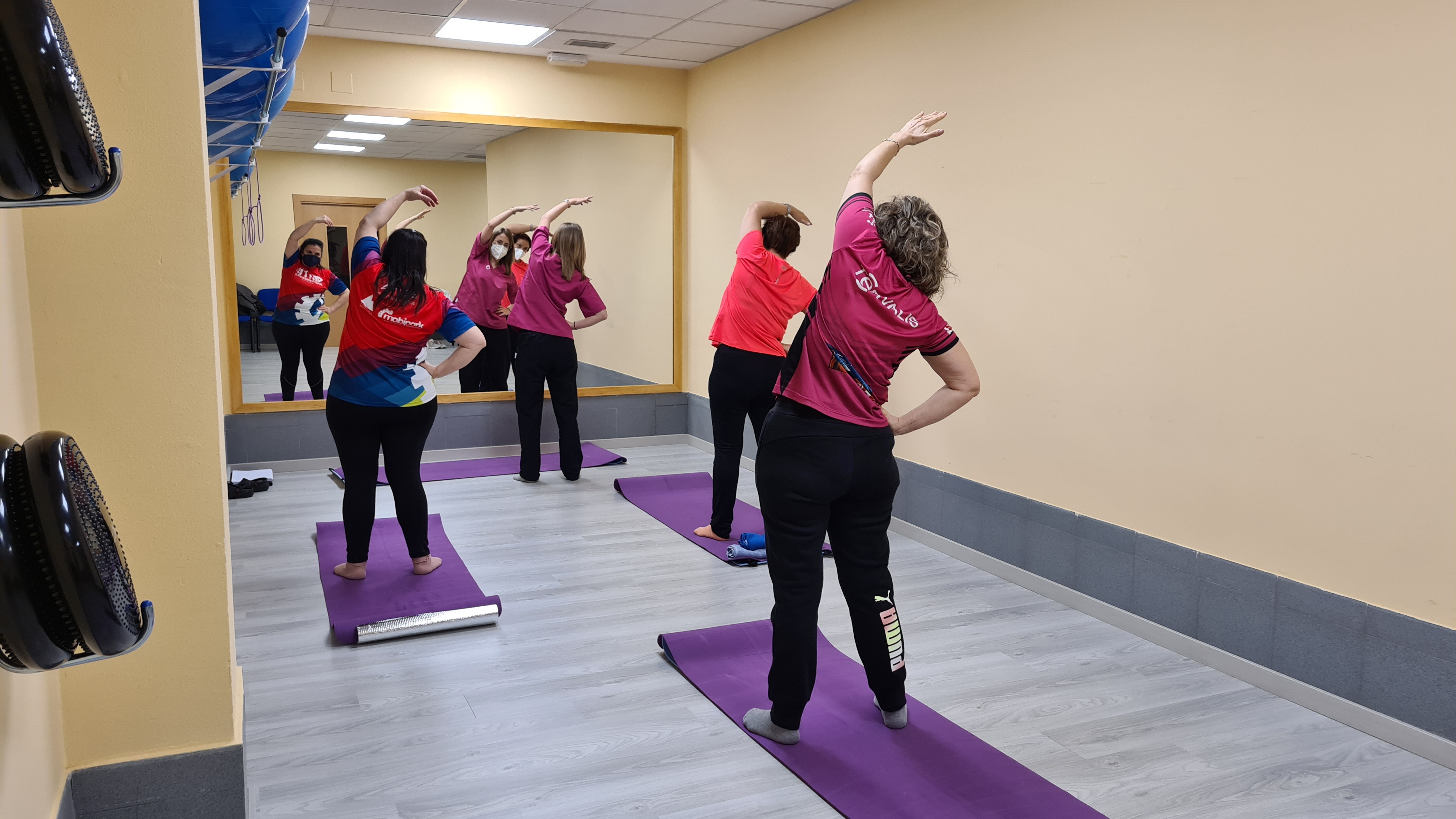 Tres mujeres y una profesora practican estiramientos frente a un espejo en una sala de gimnasio equipada