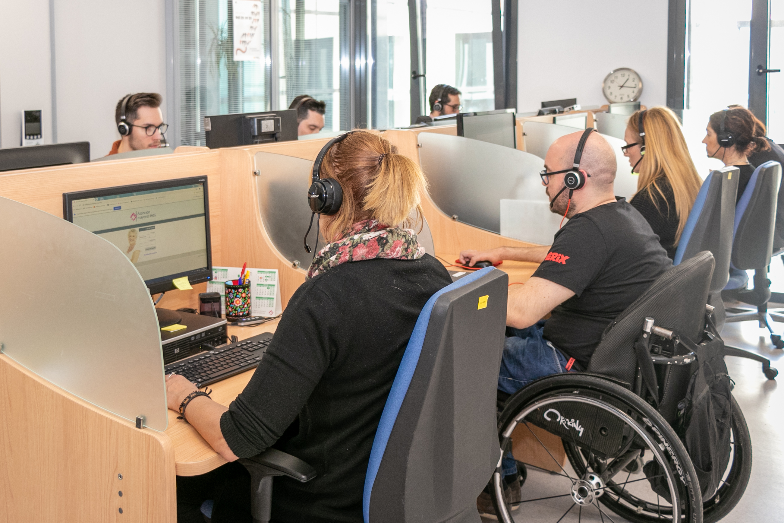 Call center con personas trabajando, algunas de ellas con discapacidad, en mesas separadas por mamparas con ordenadores y auriculares