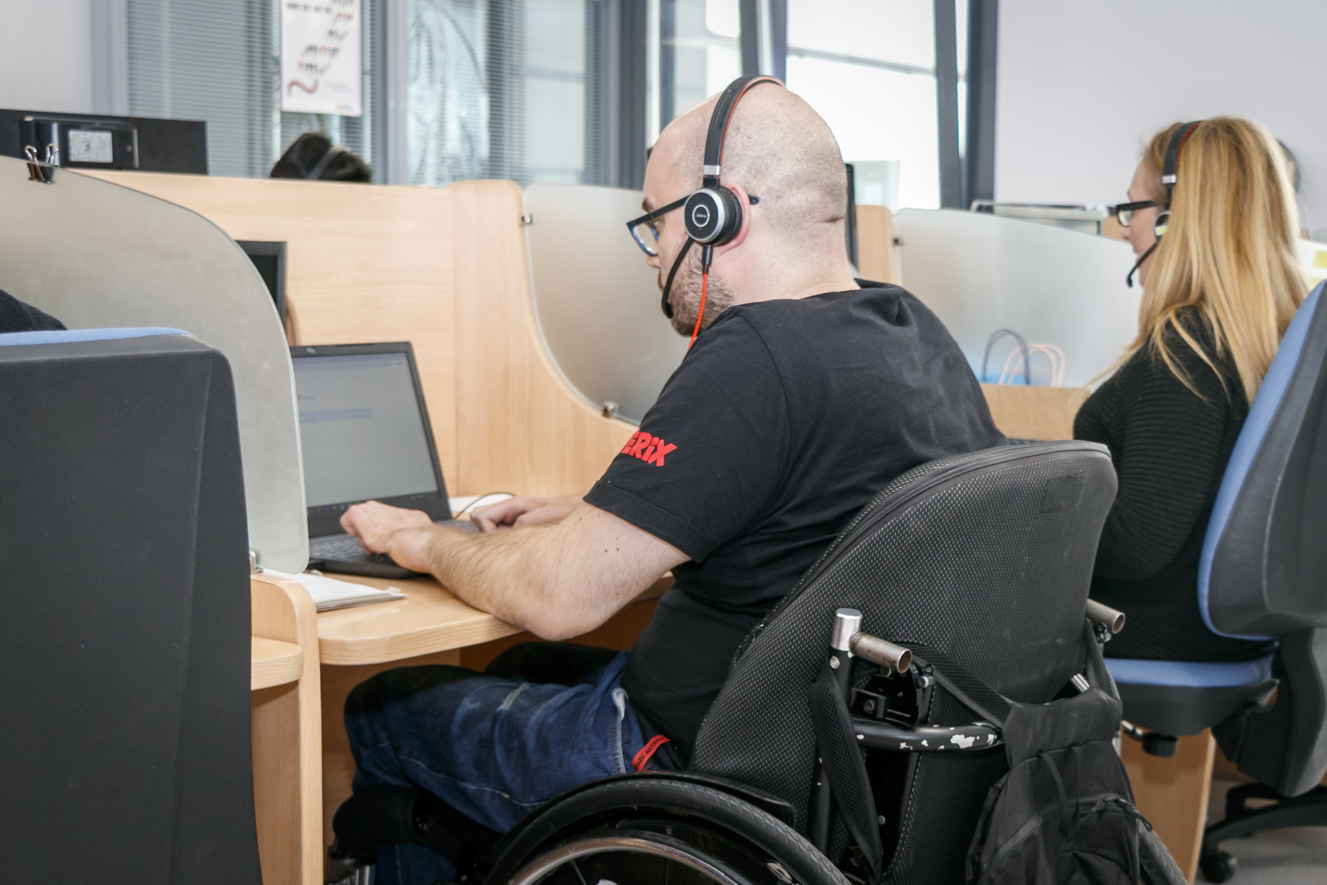Imagen de un trabajador en silla de ruedas en una sala de call center con otras personas con mesas separadas por mamparas y donde están trabajando frente a un ordenador con auriculares