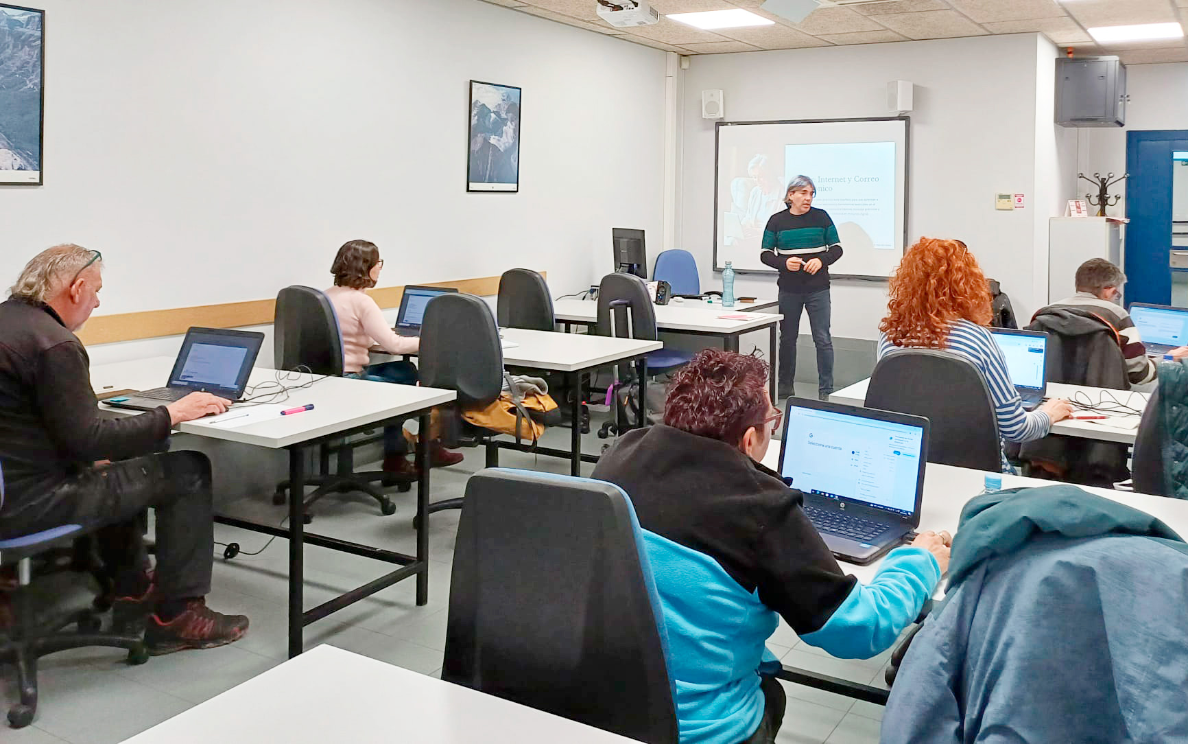 Aula de formación con varias alumnas sentadas en mesas con ordenadores y una profesora delante de una pizarra digital