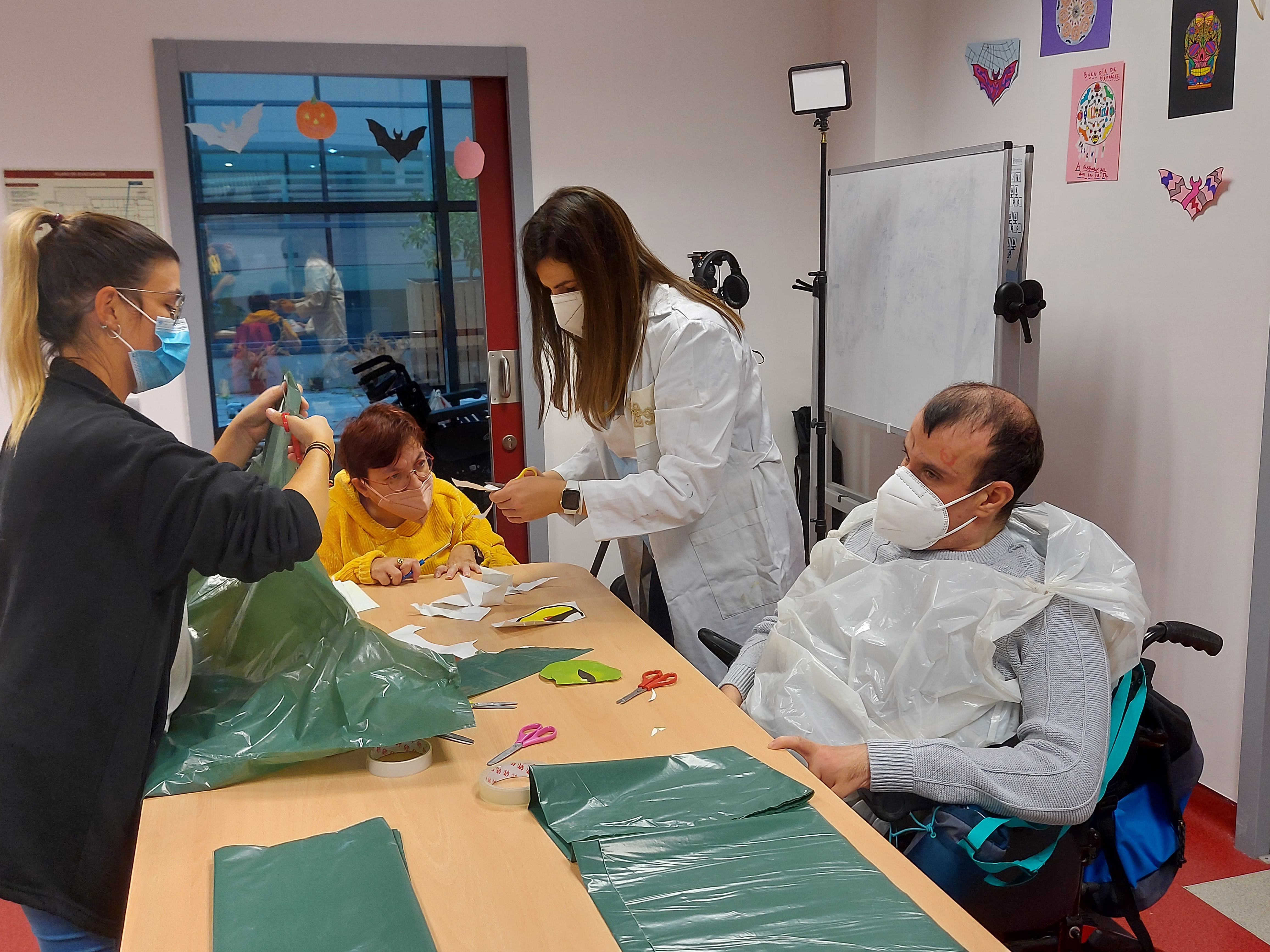 Un hombre y una mujer con discapacidad y dos voluntarias en un taller de manualidades en una mesa con pinturas, tijeras y materiales