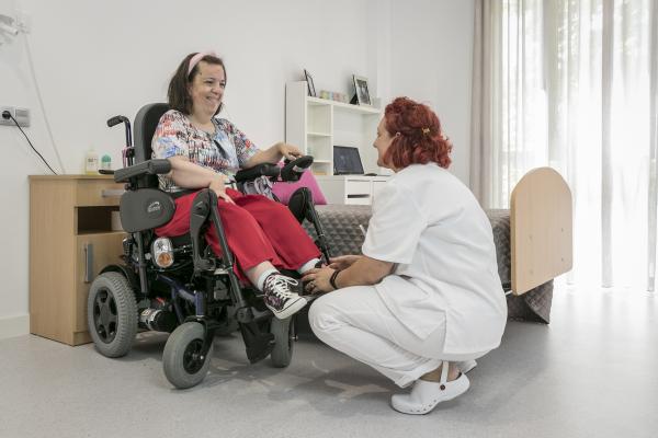 Mujer con discapacidad en silla de ruedas ayudada por una auxiliar en una habitación de una residencia