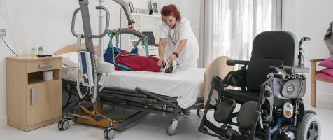 Habitación de residencia donde una auxiliar ayuda con una grúa a una mujer con discapacidad tumbada en una cama. A su lado hay una silla de ruedas automática 