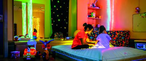 Sala de atención temprana con luces de colores y diferentes juguetes y recursos con una niña y una trabajadora sentadas en una colchoneta 