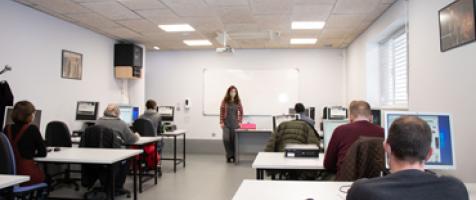 Un aula de formación con alumnado sentado ante mesas con ordenadores y una profesora de pie ante una pizarra