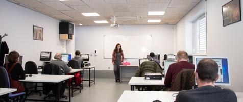 Aula de formación con varias alumnas sentadas en mesas con ordenadores y una profesora delante de una pizarra digital