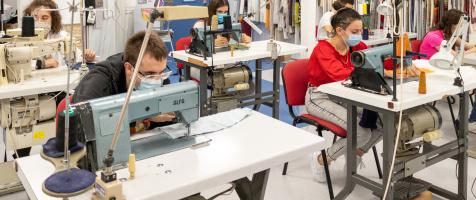 Varios chicos y chicas jóvenes en un aula de un curso textil, cada uno en una mesa con una máquina de coser y otros utensilios como telas de colores