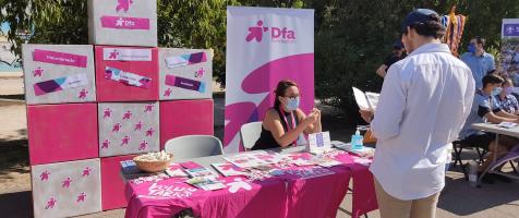 Una joven está sentada en un stand informativo decorado con material rosa mientras un joven de pie se informa con un folleto