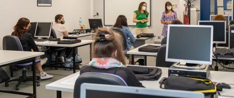 Aula de formación con varias alumnas sentadas en mesas con ordenadores y una profesora delante de una pizarra digital