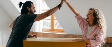 La imagen muestra a una pareja joven chocándose las manos sobre una caja cerrada en lo que parece ser una mudanza