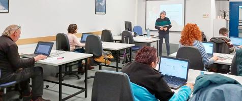 Aula de formación con varias alumnas sentadas en mesas con ordenadores y una profesora delante de una pizarra digital