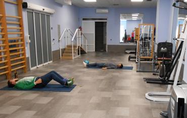 Sala de gimnasio con máquinas y equipamiento de rehabilitación con varios jóvenes haciendo ejercicios sobre una esterilla