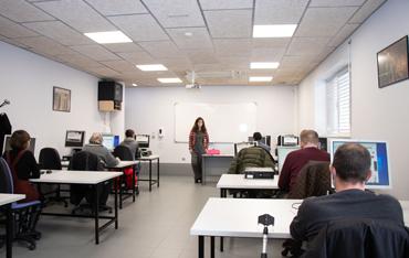 Un aula de formación con alumnado sentado ante mesas con ordenadores y una profesora de pie ante una pizarra
