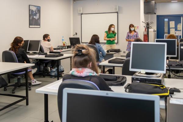 Aula con varias alumnas de espaldas y dos profesoras, con mesas, ordenadores y una pizarra digital