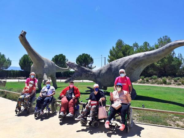 Cinco personas en silla de ruedas y dos voluntarios con camisetas rosas de la fundación posan ante las esculturas de dos dinosaurios gigantes