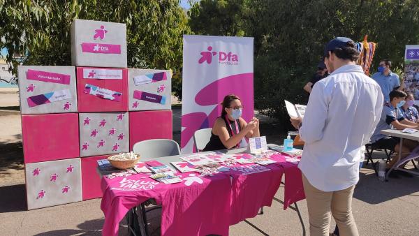 Una joven está sentada en un stand informativo decorado con material rosa mientras un joven de pie se informa con un folleto