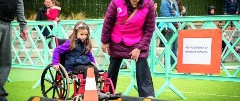 Una niña en silla de ruedas sorteando obstáculos en una yincana acompañada de una voluntaria con peto rosa